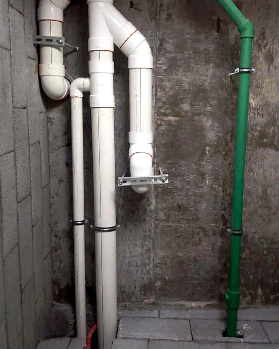 White drain risers with clamps and a green water pipe on a concrete wall