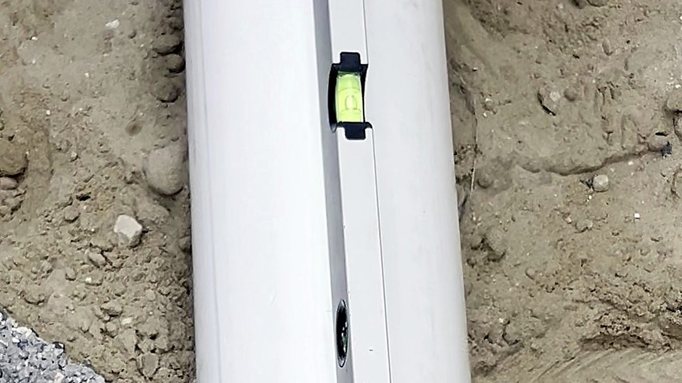 A white drain pipe laid in sand before backfilling, with a spirit level on it