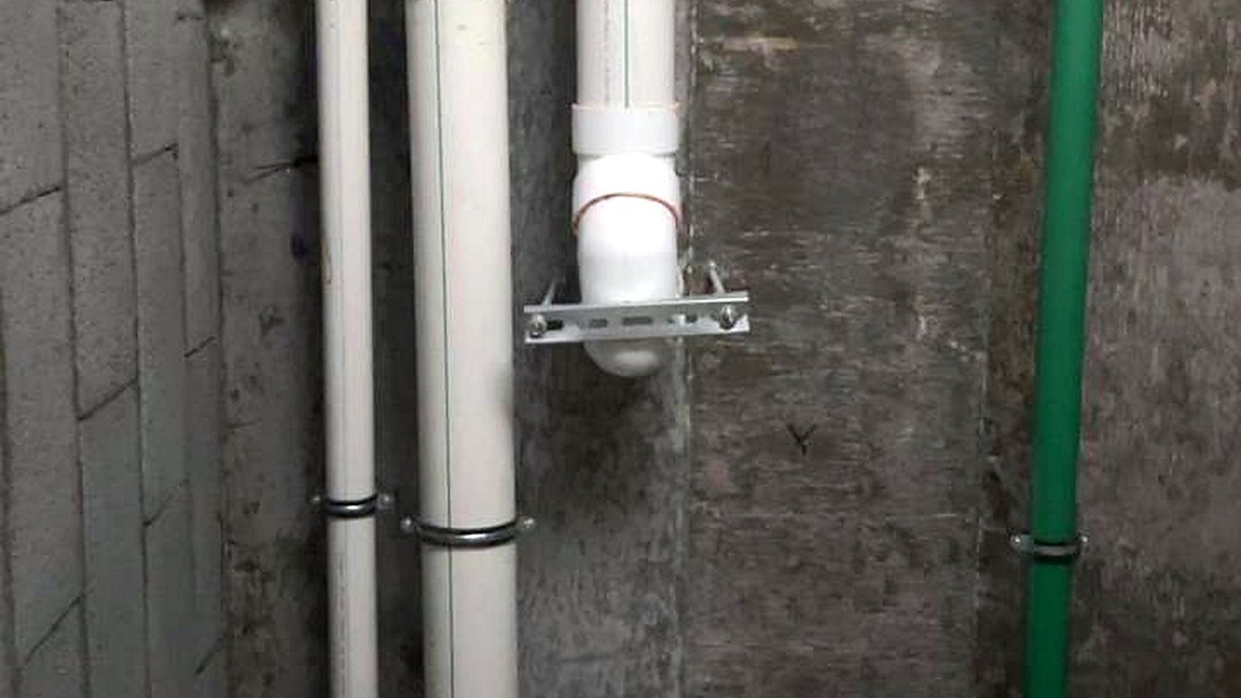 White drain risers fixed with clamps beside a green water pipe on a concrete wall
